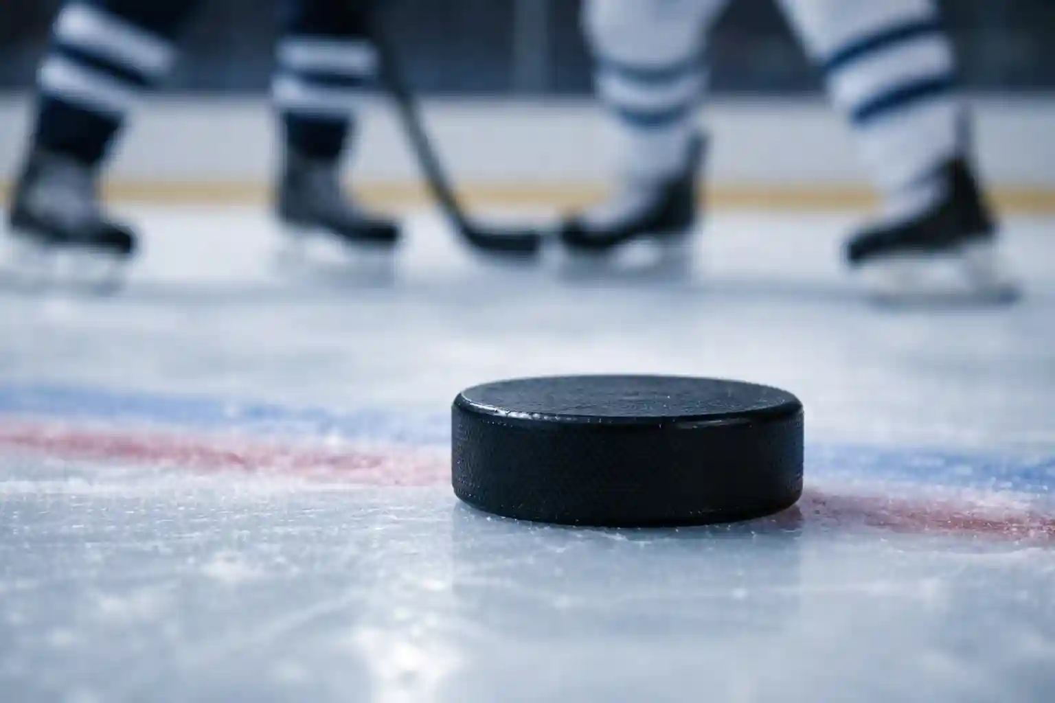 Nahaufnahme eines Eishockeypucks auf der Eisfläche mit Spielerbeinen im Hintergrund